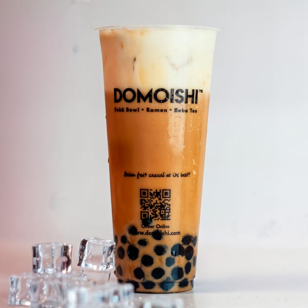 DOMOISHI boba tea with a creamy top layer, dark boba pearls, and ice cubes on a light surface.
