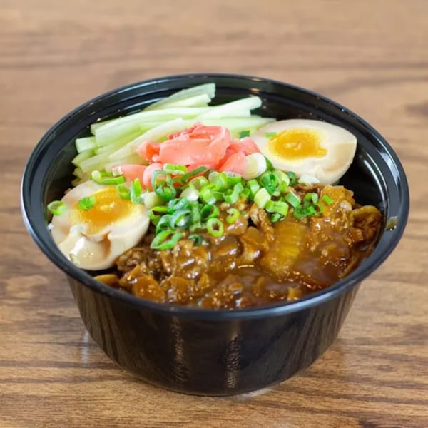 A black bowl of DOMOISHI ramen with meat, eggs, green onions, pickled ginger, and cucumbers.