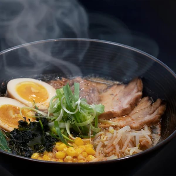 Steaming bowl of DOMOISHI ramen with pork, soft-boiled eggs, corn, seaweed, and green onions.